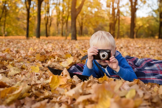 Niño rubio con cámara toma fotos. Acostado sobre una tela escocesa, hojas amarillas de otoño. Dia de otoño