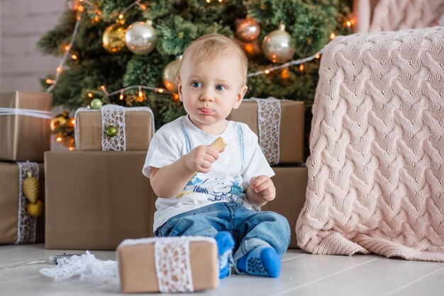 Niño rubio comiendo a un hombre de pan de jengibre junto al árbol de Navidad en casa.