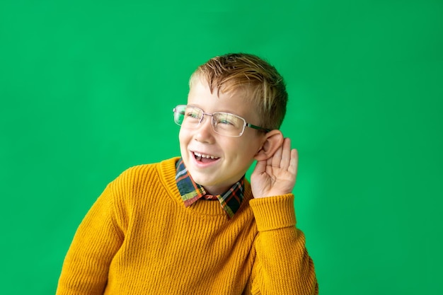 Foto un niño rubio con estilo en edad escolar tiene la mano cerca de la oreja y escucha con atención. muchacho alegre con un suéter amarillo y gafas sobre un fondo verde, aislar. regreso a la escuela, educación, aprendizaje.