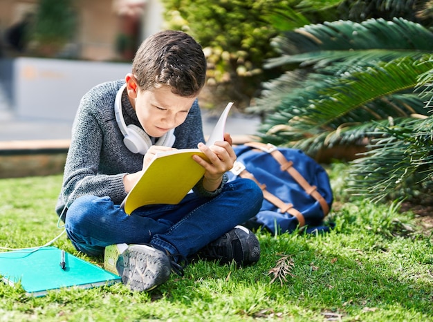 Niño rubio estudiante leyendo un libro sentado en el césped en el parque