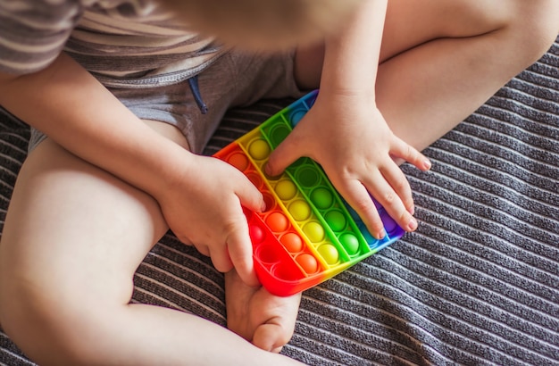 Niño rubio jugando con arco iris pop it fidget toy. Empuje el juguete sensorial bubble fidget - juguete para aliviar el estrés lavable y reutilizable. Juguete antiestrés para niño con necesidades especiales. Concepto de salud mental