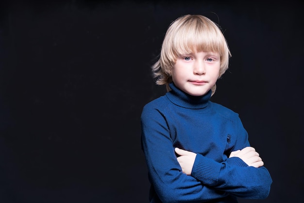 Un niño rubio de pelo largo con los brazos cruzados está parado en el estudio Fondo negro