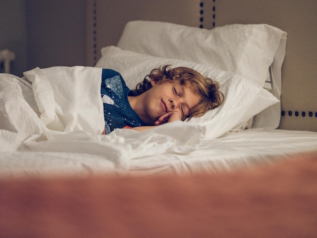 Niño rubio con pelo rizado tocando la mejilla y acostado en una cama suave mientras toma una siesta en la habitación del hotel durante el día