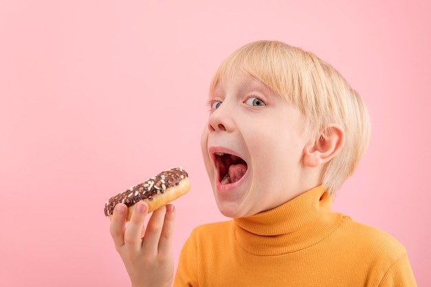 Niño rubio quiere morder donut con glaseado de chocolate Retrato de niño con donut sobre fondo rosa