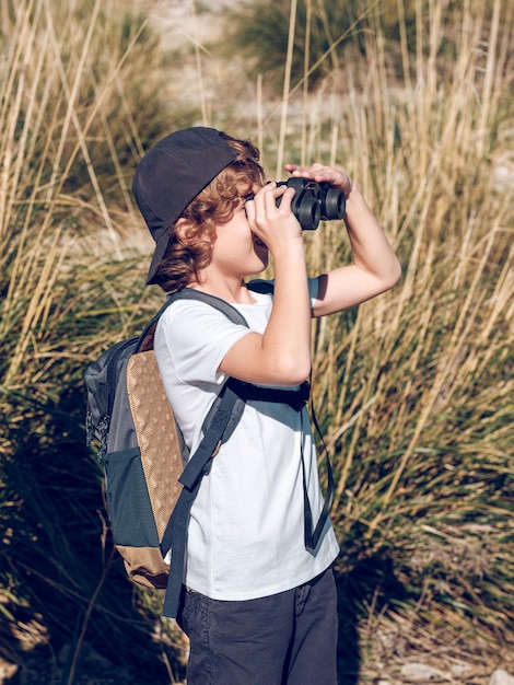 Niño rubio con ropa informal y mochila observando el campo a través de binoculares mientras se encuentra cerca de la ladera de una colina cubierta de hierba en un día soleado de fin de semana en Mallorca, España