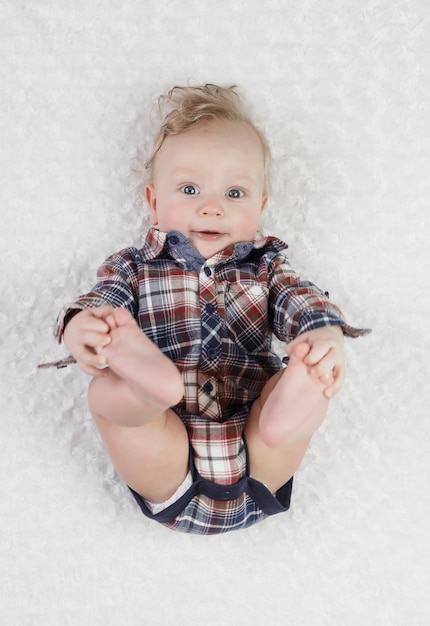 Un niño rubio de seis meses con ojos azules en una camisa a cuadros bodik yace jugando con sus piernas, sonriendo lindo,