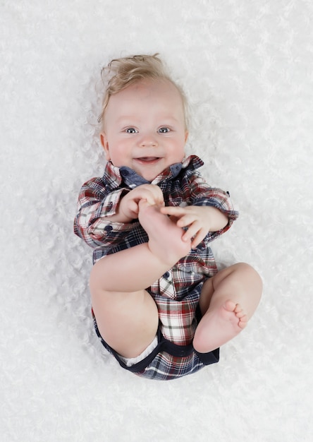 Un niño rubio de seis meses con ojos azules en una camisa a cuadros bodik yace jugando con sus piernas, sonriendo lindo,