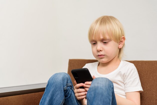 Niño rubio sentado en el sofá y se ve en el teléfono inteligente. Apps para niños