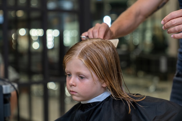 Un niño rubio se sienta en una silla en una peluquería y un estilista se peina antes de cortar