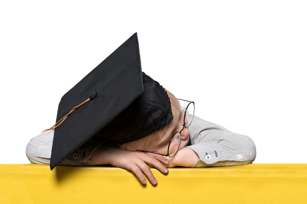 Niño rubio con un sombrero académico y gafas dormido sobre la mesa.