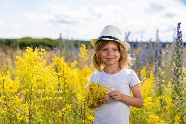 Un niño rubio con un sombrero de paja sonriendo mientras está de pie en un campo con flores que crecen en él