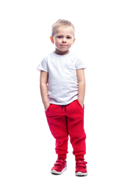 Un niño rubio sonriente se para con las manos en los bolsillos Un niño con pantalones rojos y una camiseta blanca Aislado sobre fondo blanco Vertical de altura completa