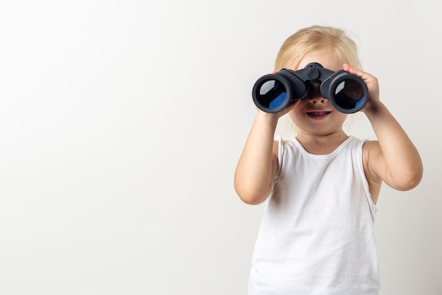 Foto niño rubio sonriente mira a través de binoculares sobre un fondo claro en el estudio.