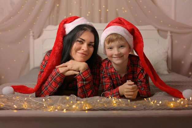 Un niño rubio y su madre con un gorro de Papá Noel en pijama de Navidad están acostados en la cama