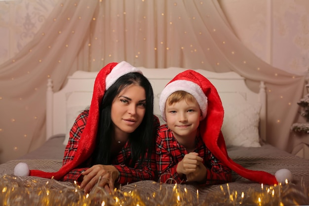 Un niño rubio y su madre con un gorro de Papá Noel en pijama de Navidad están acostados en la cama