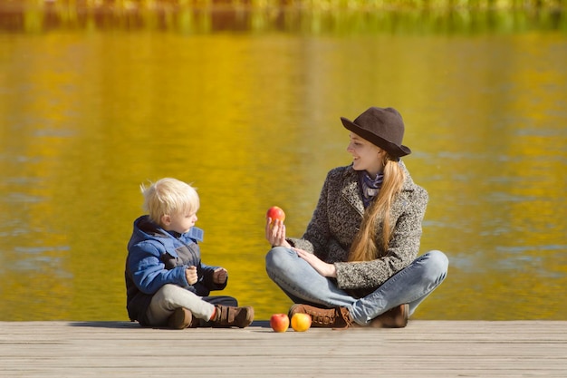 Niño rubio con su madre sentada en el muelle y jugando con manzanas Día soleado de otoño