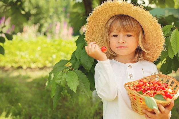 un niño rubio con un traje blanco de lino y un sombrero de paja sostiene una canasta con cerezas