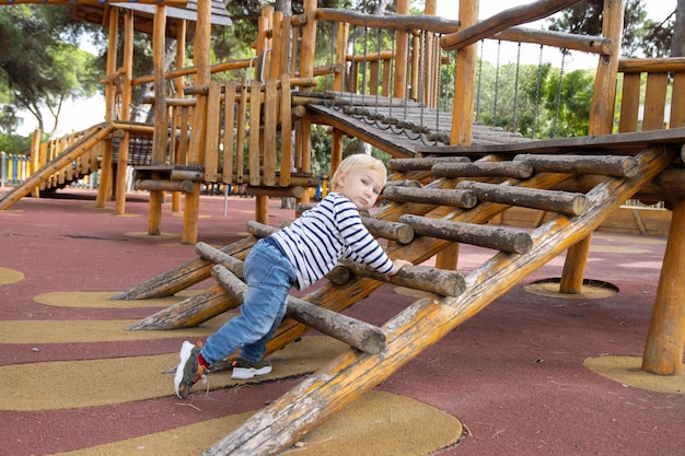 Un niño rubio trepando por barras de madera en el patio de recreo mirando a la cámara