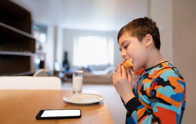 Niño sano desayunando Niño feliz Niño usando un teléfono móvil mirando leyendo o jugando mientras come comida School Kid croissant y leche antes de ir a la escuela por la mañana