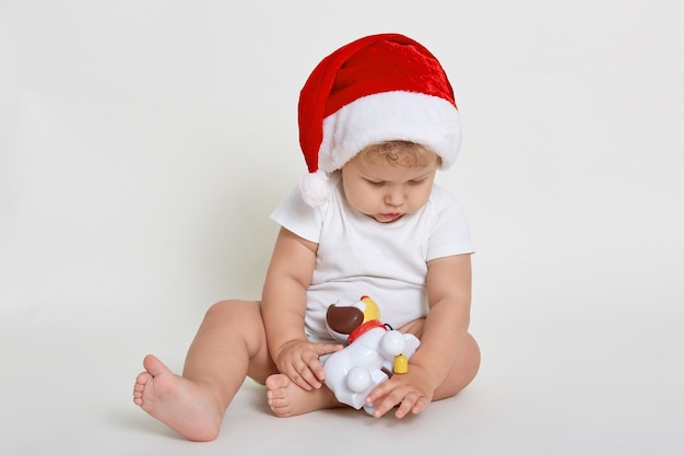 Niño de Santa jugando con juguete de niebla aislado sobre una pared blanca, bebé descalzo en mono mirando concentrado en juguete, bebé mirando hacia abajo, lindo bebé en Nochebuena.