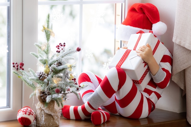 Niño sentado en el alféizar de la ventana Niño feliz vistiendo pijamas de Navidad