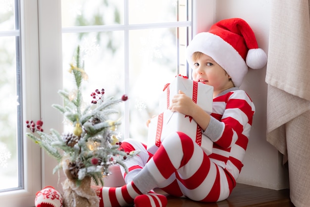 Niño sentado en el alféizar de la ventana Niño feliz vistiendo pijamas de Navidad