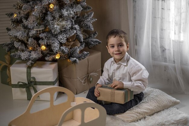 Foto el niño está sentado en la alfombra cerca del árbol de navidad sosteniendo un regalo y jugando con una máquina de escribir de madera