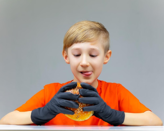 Un niño sentado en una mesa con guantes negros desechables sostiene frente a él una enorme hamburguesa rociada con semillas de sésamo, un niño que mira una hamburguesa se lame los labios