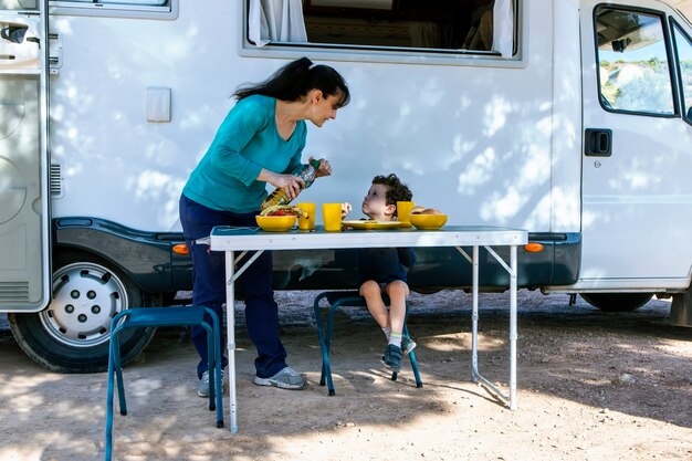 Niño sentado en la mesa junto a la caravana mientras su madre sirve bocadillos en un día de campamento