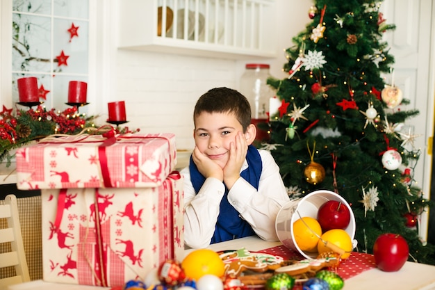 niño sentado en la mesa con regalos cerca y el árbol de navidad