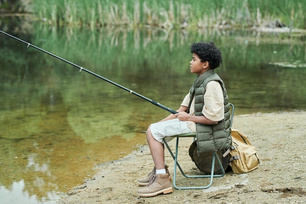 Niño sentado en una silla con caña de pescar y pescando cerca del lago