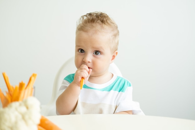 Niño sentado en una silla para niños comiendo rodajas de zanahoria sobre fondo blanco.