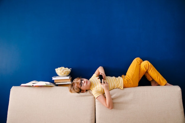 Niño sentado en el sofá, comiendo palomitas de maíz y jugando con gamepad durante su lección en línea en casa, distancia social durante la cuarentena, autoaislamiento, concepto de educación en línea