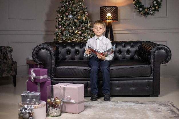 Foto el niño está sentado en el sofá y leyendo un libro en la víspera de año nuevo. detrás de él hay un árbol de navidad decorado con juguetes y luces frente a muchos regalos.