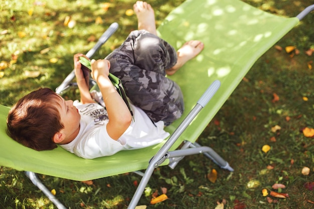 Niño sentado con una tableta en el jardín el niño se acuesta en una tumbona y juega en la tableta niño con un gadget