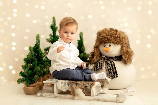 Niño sentado en un trineo cerca del árbol de Navidad y el muñeco de nieve
