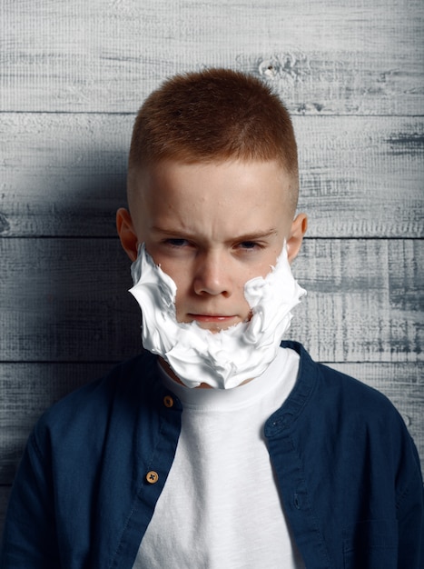 Niño serio con espuma de afeitar en la cara en estudio. Niño aislado sobre fondo de madera, emoción infantil, sesión de fotos de colegial