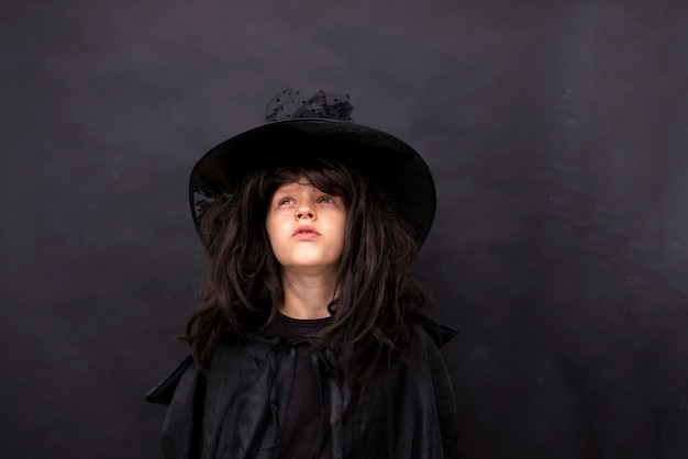 Niño serio de Halloween con fondo negro mira hacia arriba Niño con traje de hechicero negro con cabello largo Truco de bruja misterioso o trato alegre Niño con traje de mago Espacio de copia