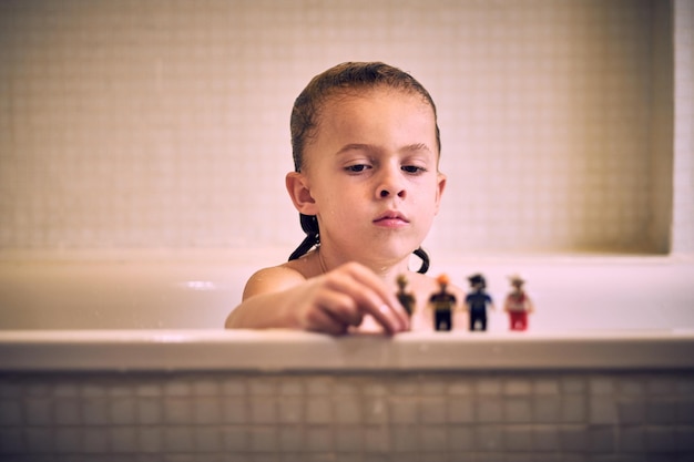 Niño serio con el pelo mojado jugando con juguetes mientras está sentado en una bañera blanca durante la rutina diaria de higiene en un baño ligero