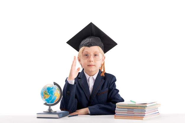 Niño serio de primer grado con sombrero de estudiantes levantando la mano junto a cuadernos y globo Concepto de estudio y educación