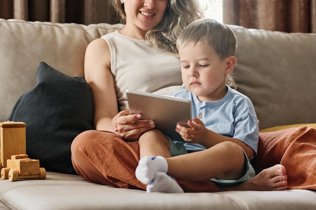 Niño serio en ropa casual mirando la pantalla de la tableta sostenida por su madre