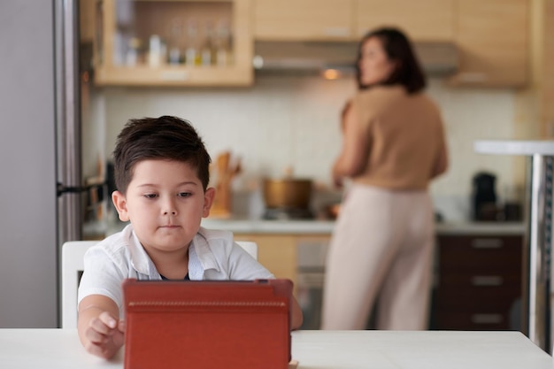 Un niño serio viendo videos educativos en una tableta cuando su madre cocina la cena en b