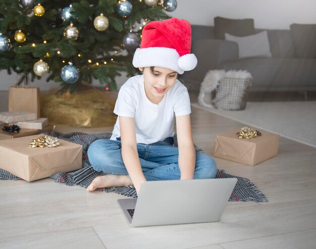 Un niño se sienta cerca de un árbol de Navidad con una computadora portátil. Árbol de Navidad en una gran sala de estar.