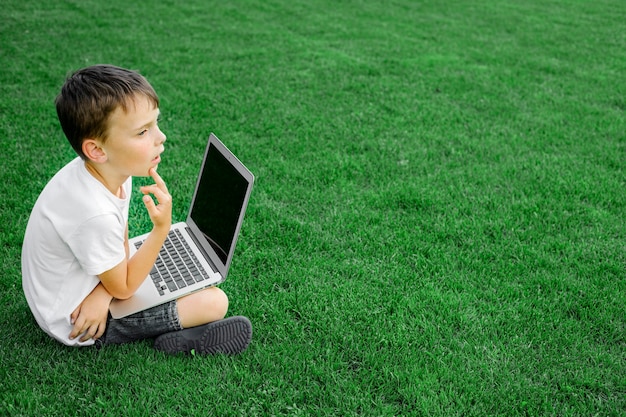 Un niño se sienta en el césped y estudia con una computadora portátil en línea, disfrutando de la naturaleza