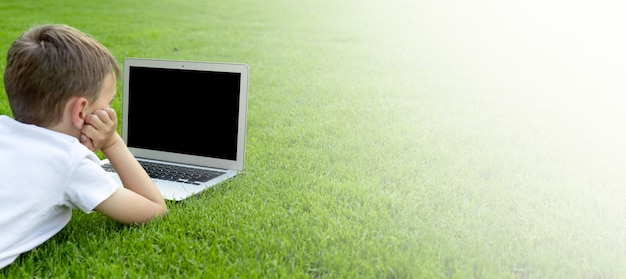 Un niño se sienta en el césped y estudia con una computadora portátil en línea disfrutando de la naturaleza