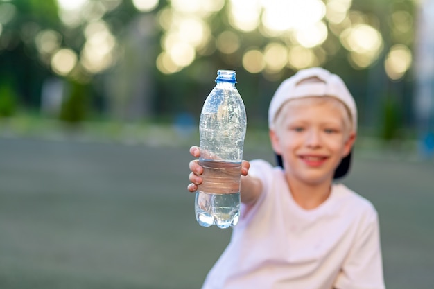Un niño se sienta en un césped verde en un campo de fútbol y sostiene una botella de agua. Concéntrese en la botella de agua.