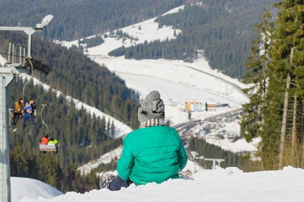 Niño se sienta en una colina cubierta de nieve en el fondo del teleférico, bosque de pinos y montañas