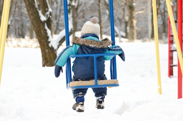 Niño se sienta en un columpio buen tiempo día de invierno Vista trasera