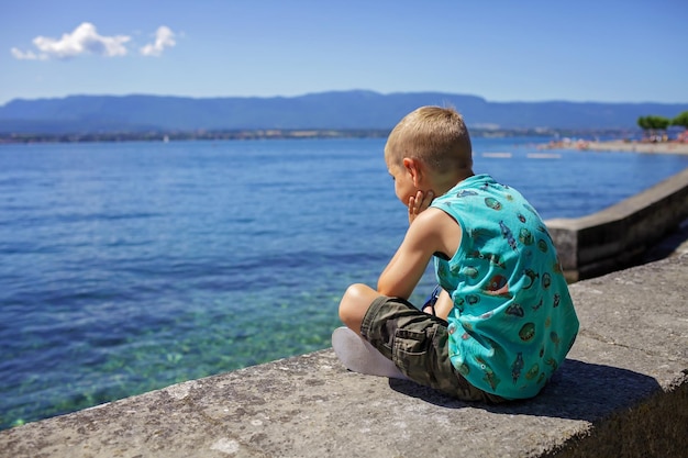 El niño se sienta en el muelle del lago de ginebra y mira el agua y las montañas, vacaciones y viajes de verano.