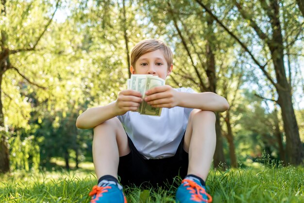 Un niño se sienta en un parque en el césped sosteniendo una pila de billetes de un dólar frente a él.